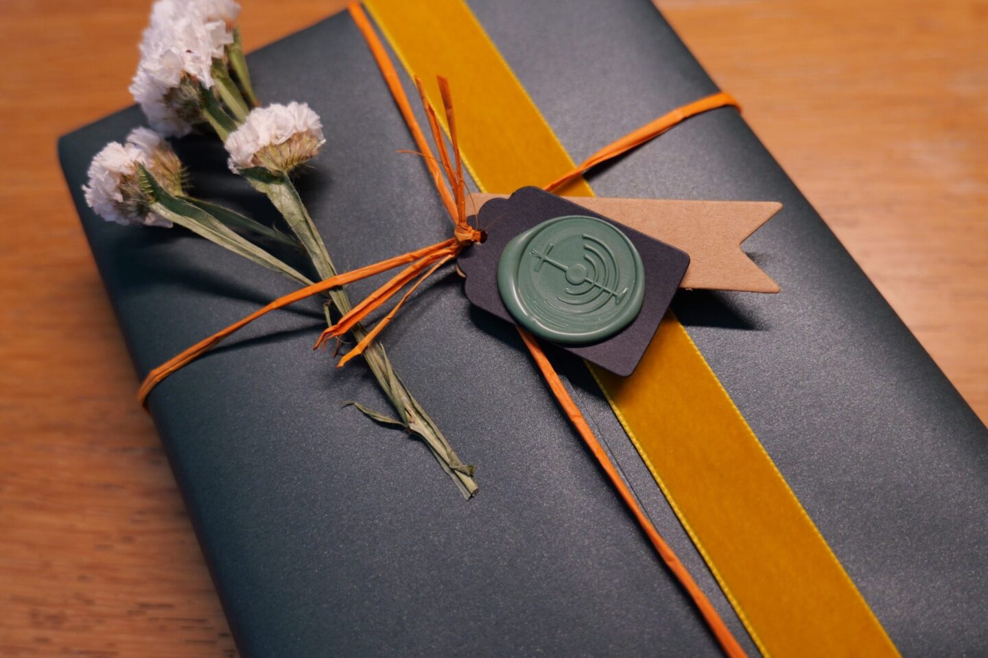 Dark green wrapped gift with orange twine, yellow ribbon, dried flowers, and wax seal
