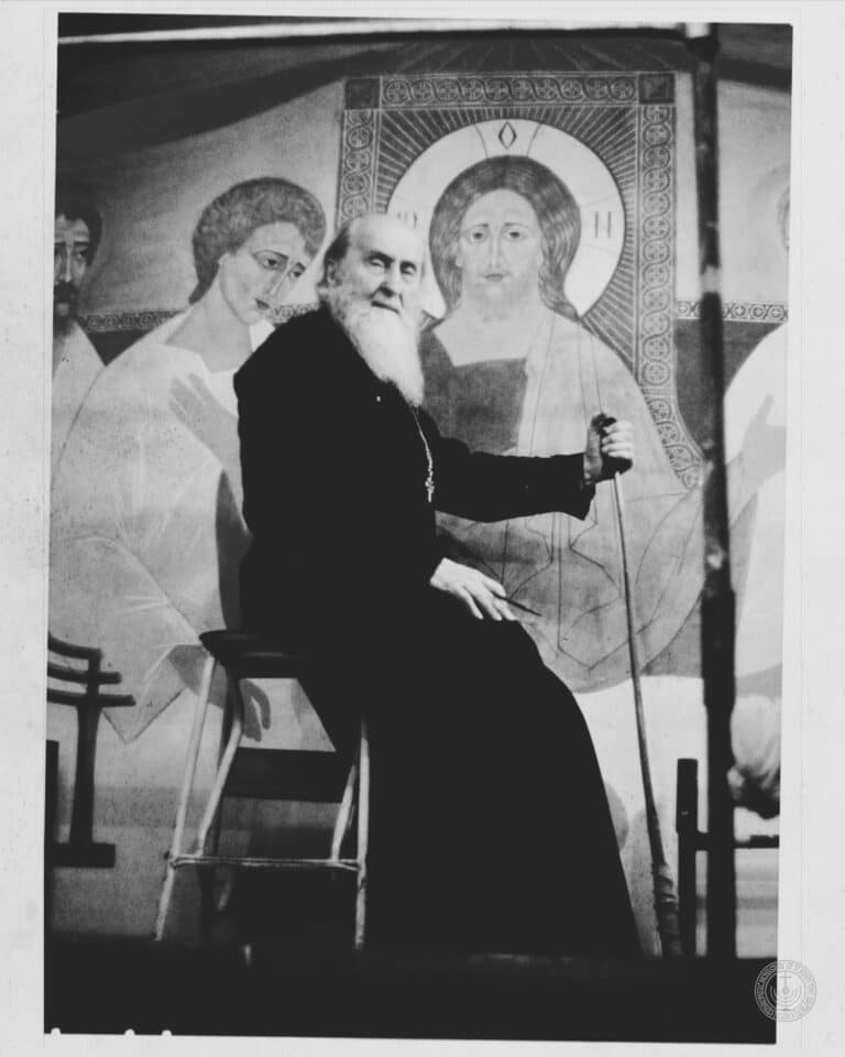 Elderly Orthodox priest with long white beard sitting before a religious mural.